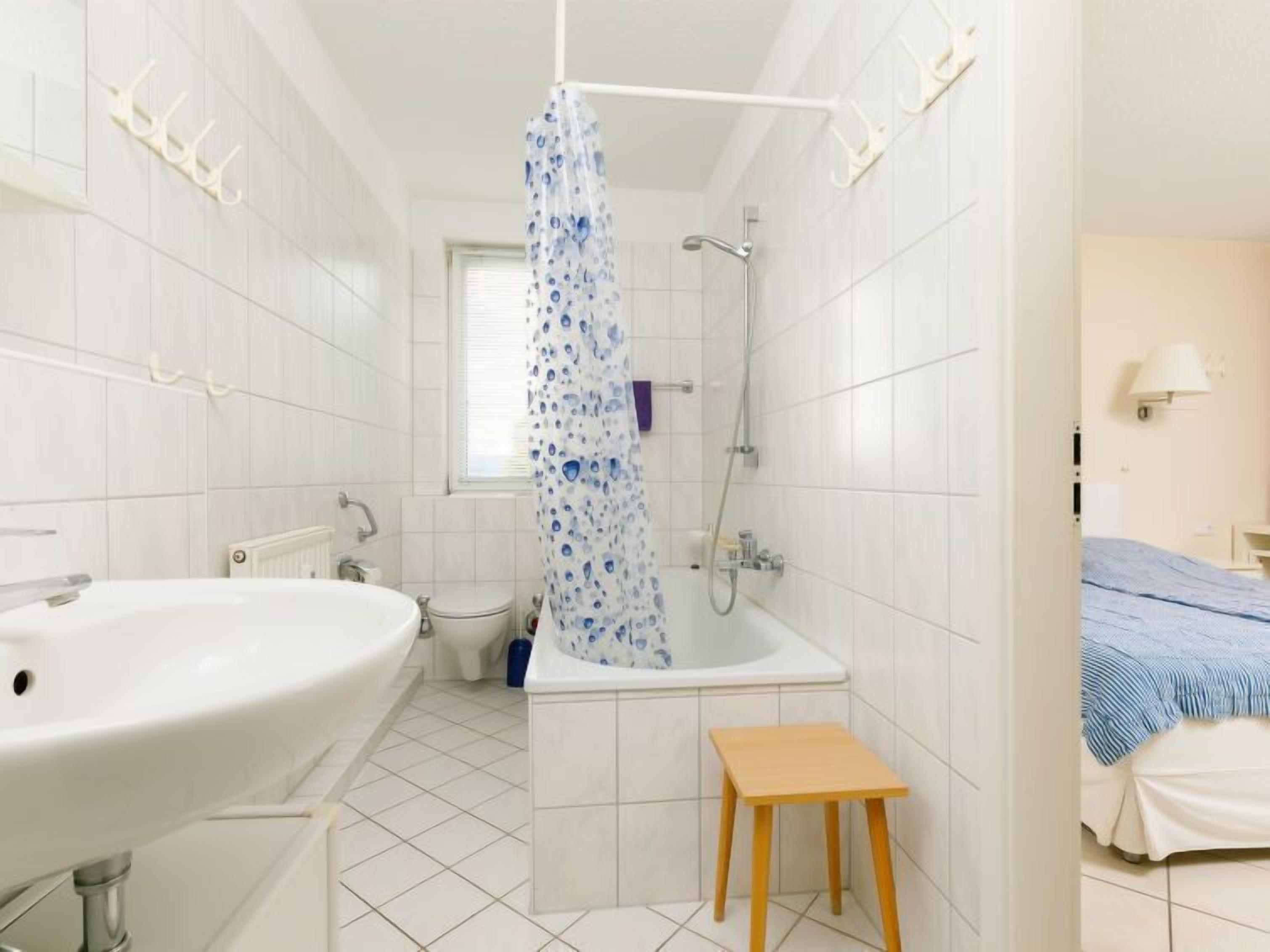Un baño con bañera, inodoro y lavabo. Hay una cortina de ducha con un estampado azul. Un taburete de madera pequeño está colocado junto a la bañera.