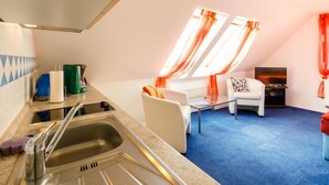 Ett kompakt bostadsutrymme med ett litet kök, en sittgrupp med TV och ett fönster med gardiner.