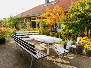 An outdoor patio with a wooden bench and a table, surrounded by lush greenery and a brick building in the background. - Tauchmann II by Interhome (Koserow)
