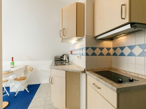 Een kleine keuken met een gasfornuis, een broodrooster en een spoelbak. Er ligt een blauw tapijt en is een klein eethoekje met stoelen.