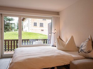 A bedroom with a bed, a nightstand, and a window with a view of a building and greenery outside. - Chesa Palüdin 10 - Pontresina by Interhome (Pontresina)