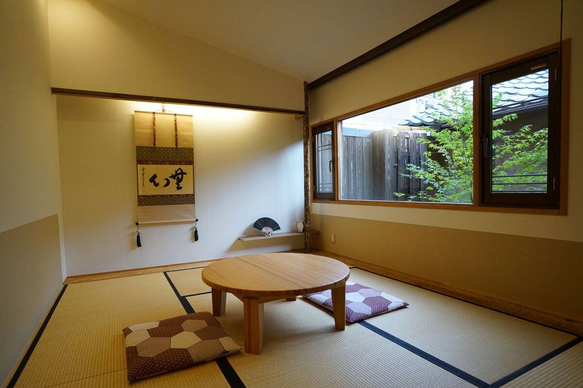 Een kamer met tatami matten, een ronde tafel, kussens, een hangende scroll en een raam met uitzicht op groen.