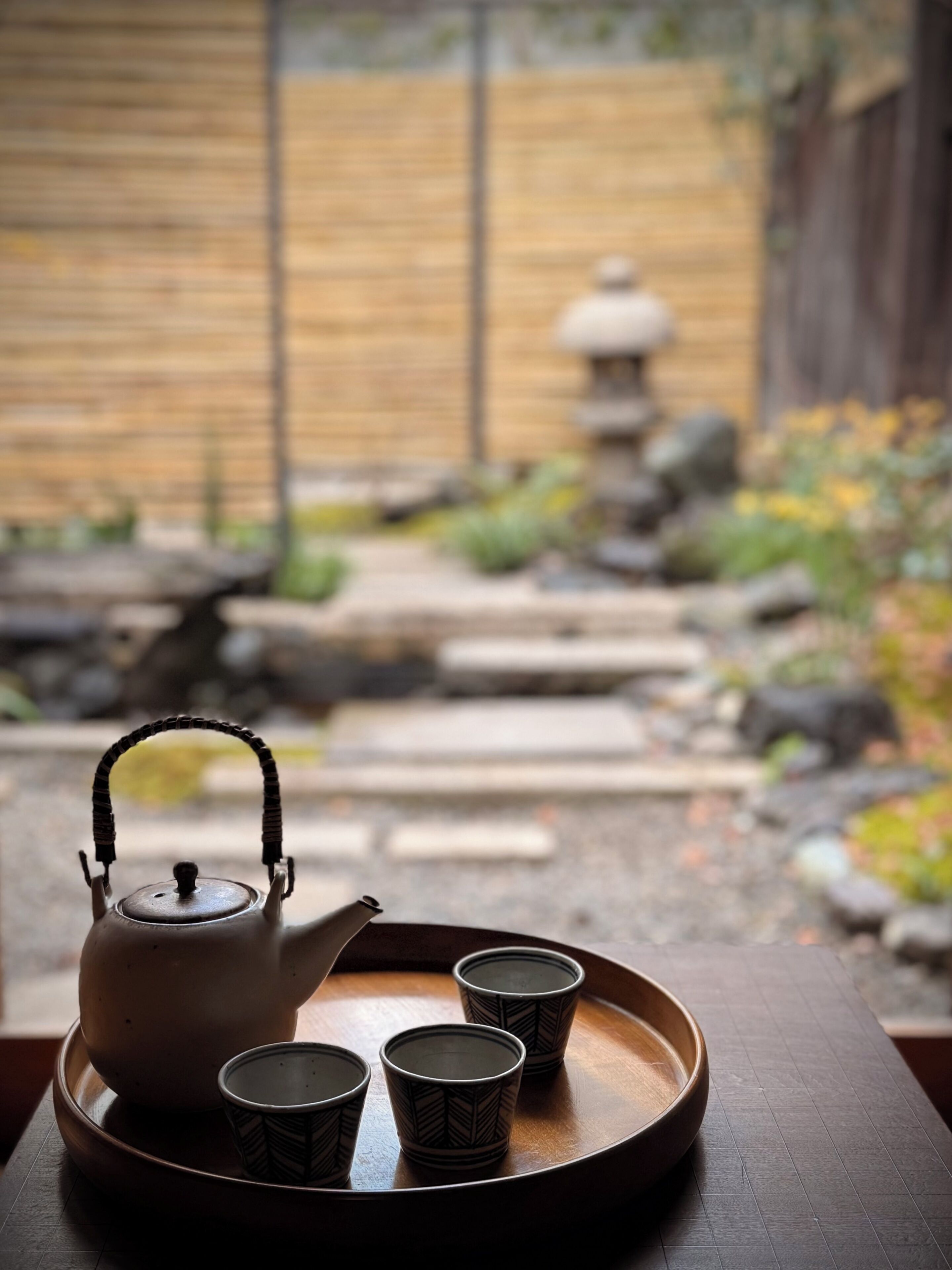 Un service à thé traditionnel, posé sur un plateau, sur une table en bois, avec un jardin flou en arrière-plan.
