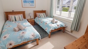 Une chambre avec deux lits simples, une table de chevet, un banc en bois, une fenêtre avec des rideaux et un tableau encadré au mur.