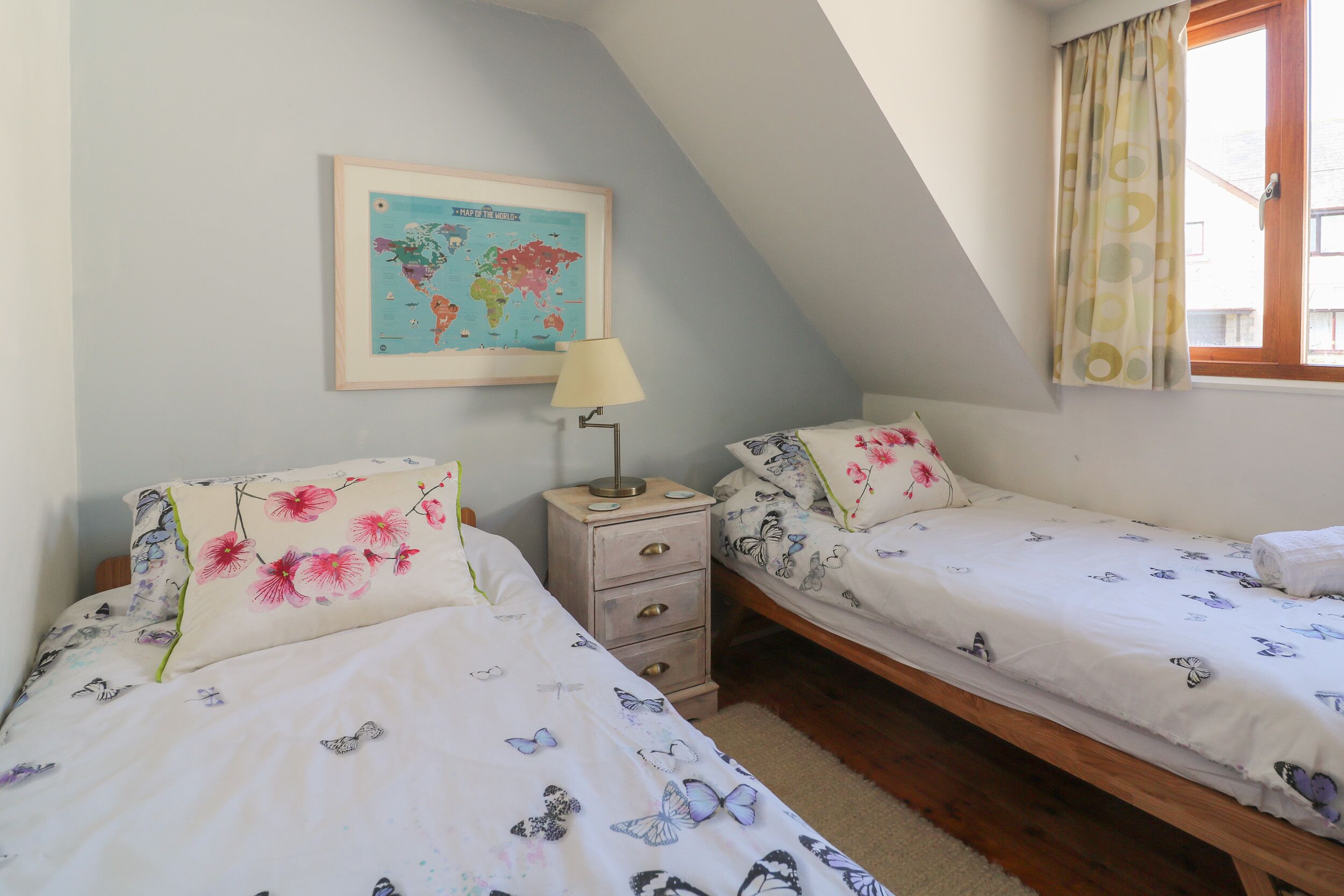 Un dormitorio con dos camas individuales, una mesita de noche, una lámpara, un mapa del mundo en la pared y una ventana con cortinas.