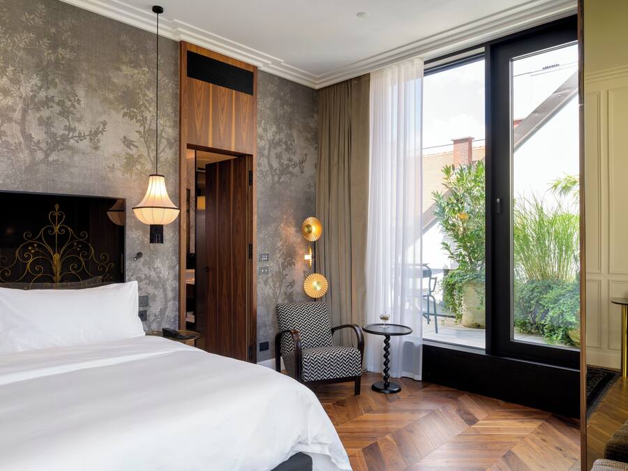 A hotel room with a large bed, a wooden wardrobe, a small table with a lamp, and a view of greenery outside.