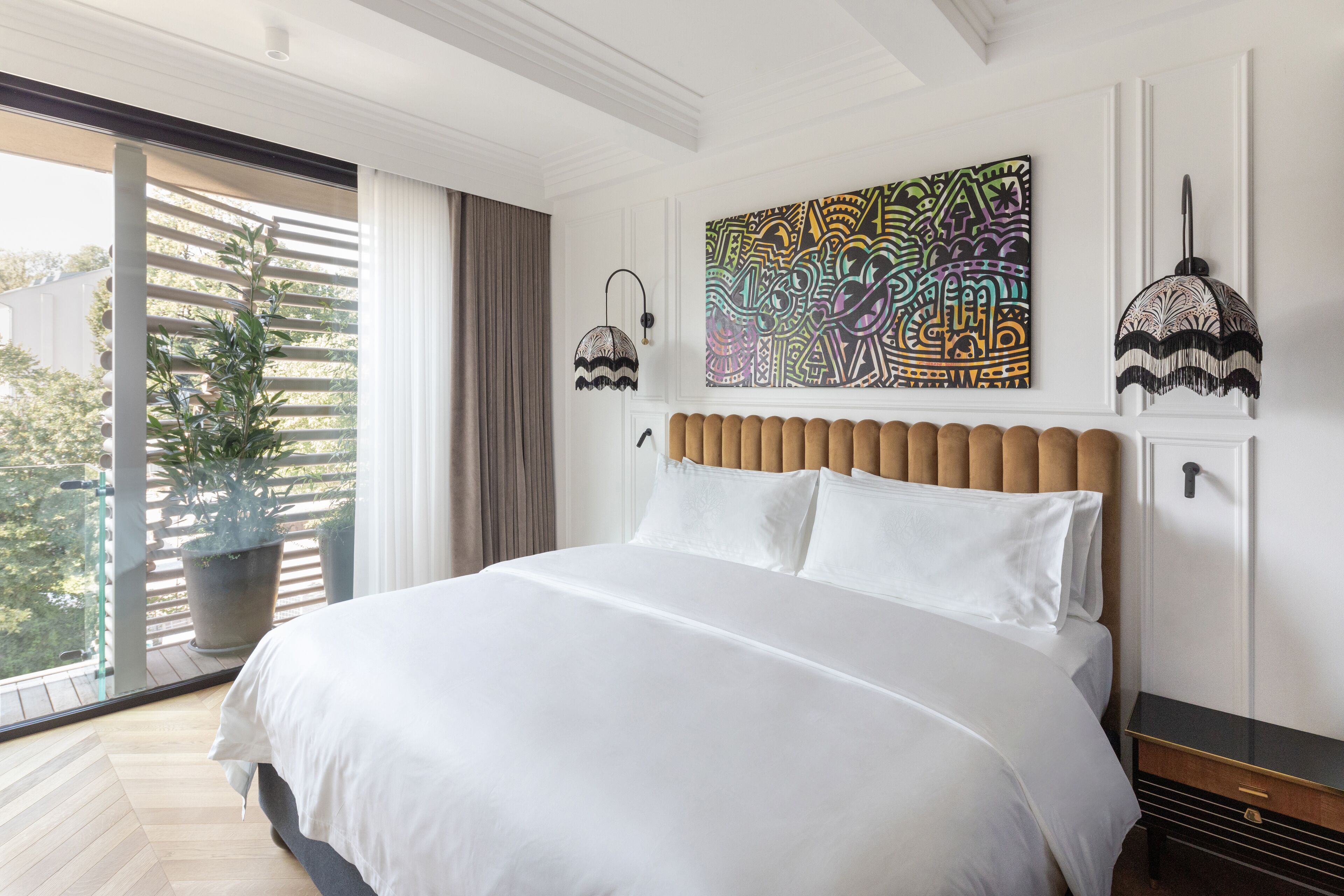 A modern bedroom with a large bed, a view of greenery outside, and a colorful abstract painting on the wall.