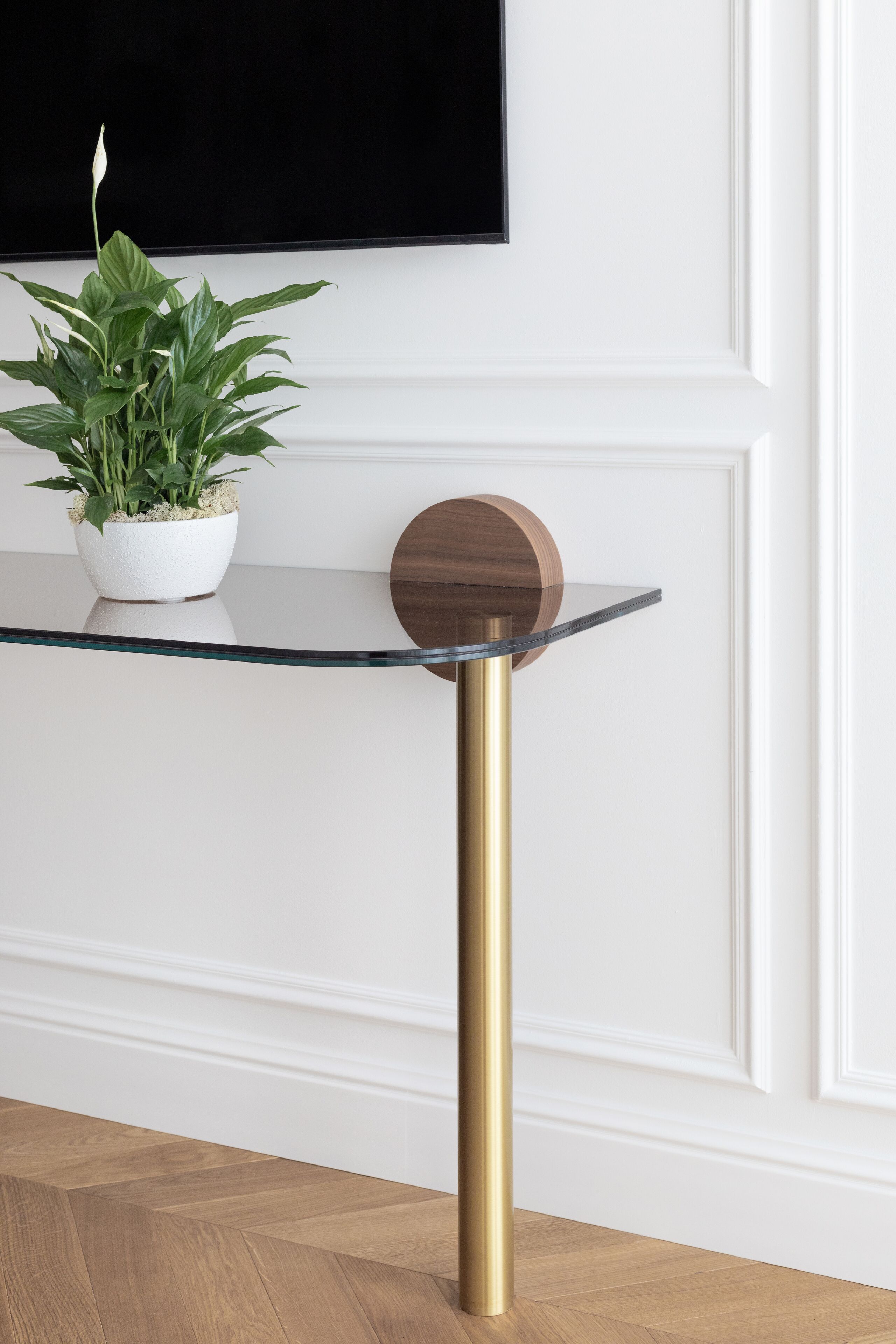 A modern table with a glass top and a gold pole, accompanied by a potted plant and a wooden sphere.