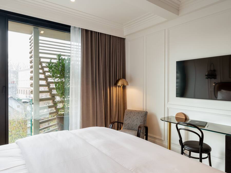 A hotel room with a large bed, a desk with a chair, a TV mounted on the wall, and a view of the city through a large window.