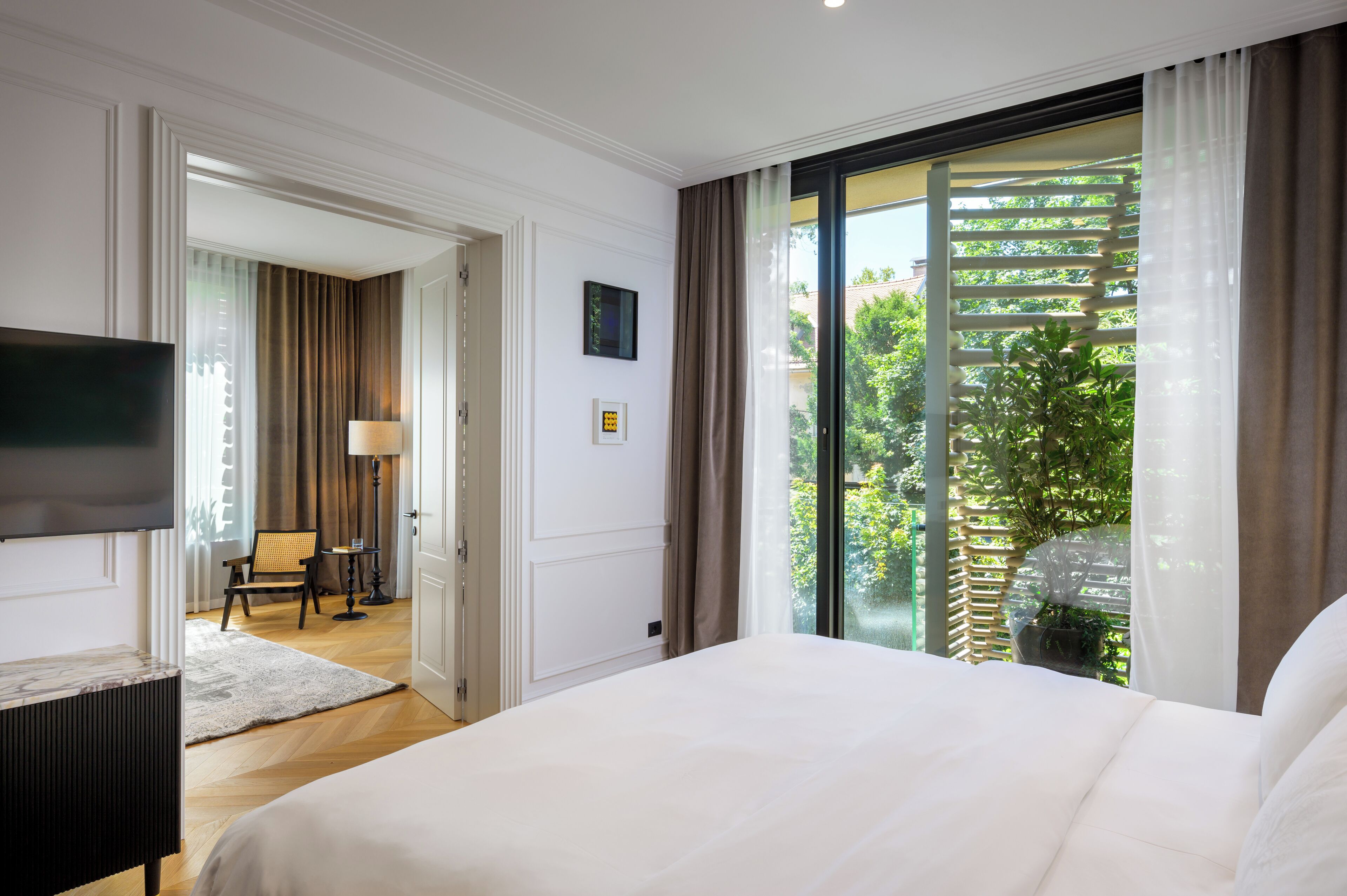 A hotel room with a large bed, a flat-screen TV, a desk, and a view of greenery through a sliding door.