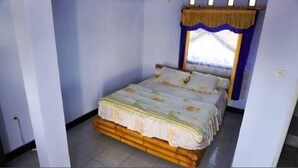 A single bed with a wooden frame and a floral bedspread in a room with light blue walls and a window with a blue curtain. - Hans Yoga Homestay (Kuta)
