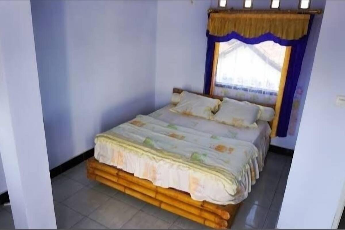 A single bed with a wooden frame and a floral bedspread in a room with light blue walls and a window with a blue curtain.