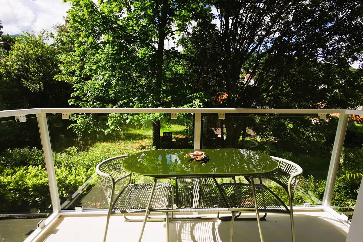 Un balcon doté d’une table en verre et de chaises en métal, donnant sur un jardin luxuriant.