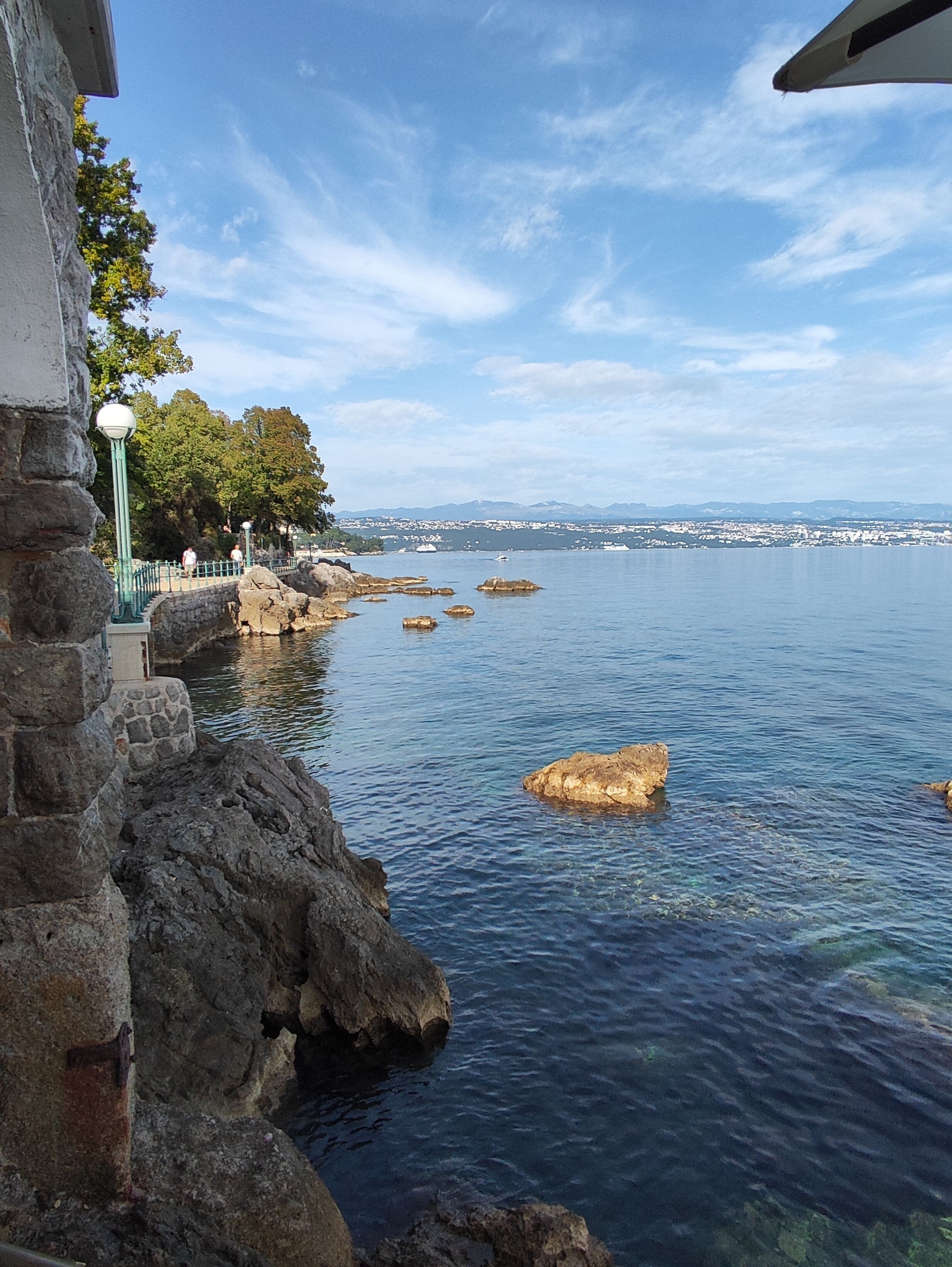 A coastal view with clear water, rocky shoreline, and distant cityscape.