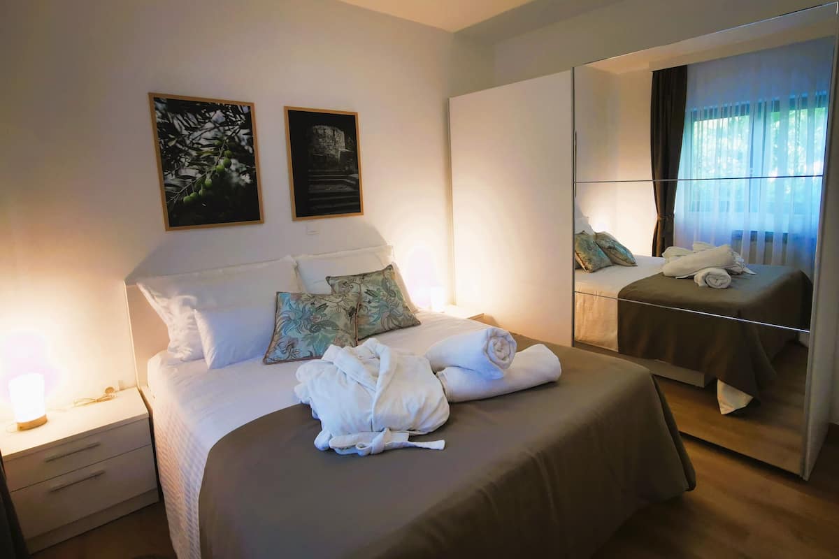 Un lit bien fait, avec des oreillers et une serviette pliée, une table de chevet avec une lampe, et deux tableaux encadrés accrochés au mur.