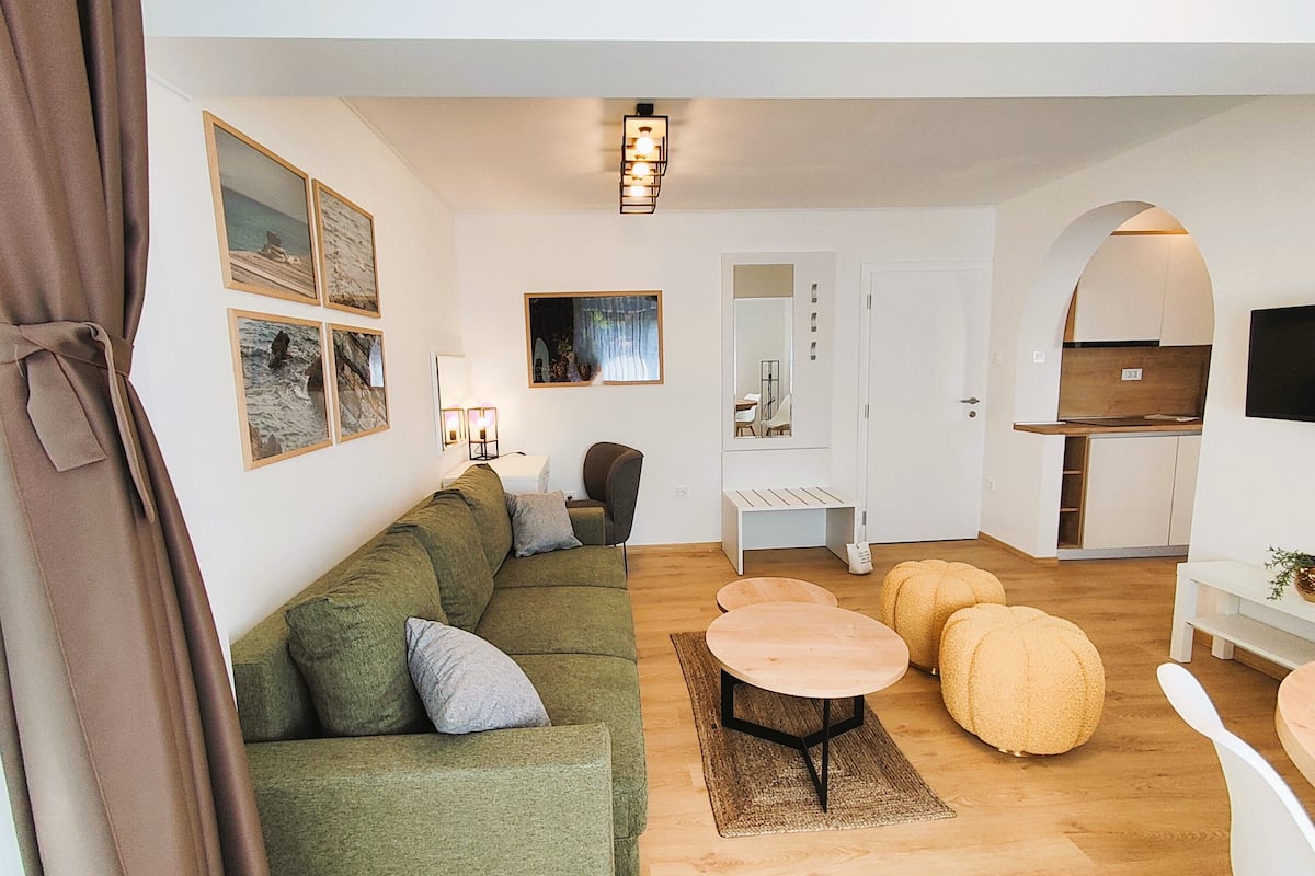 Un salon moderne avec un canapé vert, une table basse ronde en bois et deux poufs. On y trouve également un coin cuisine avec un téléviseur et une table à manger.