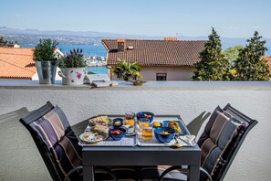 Ein Esstisch mit gedecktem Tisch, Stühlen und Blick auf das Meer sowie Gebäude im Hintergrund.