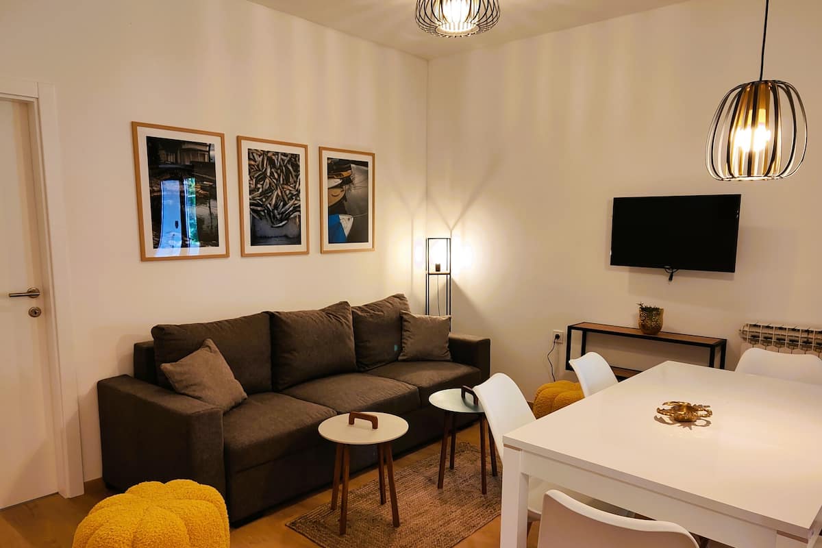 Un salon moderne comprenant un canapé, une petite table ronde, une table à manger avec des chaises, un téléviseur à écran plat fixé au mur et trois œuvres d’art encadrées accrochées au mur.