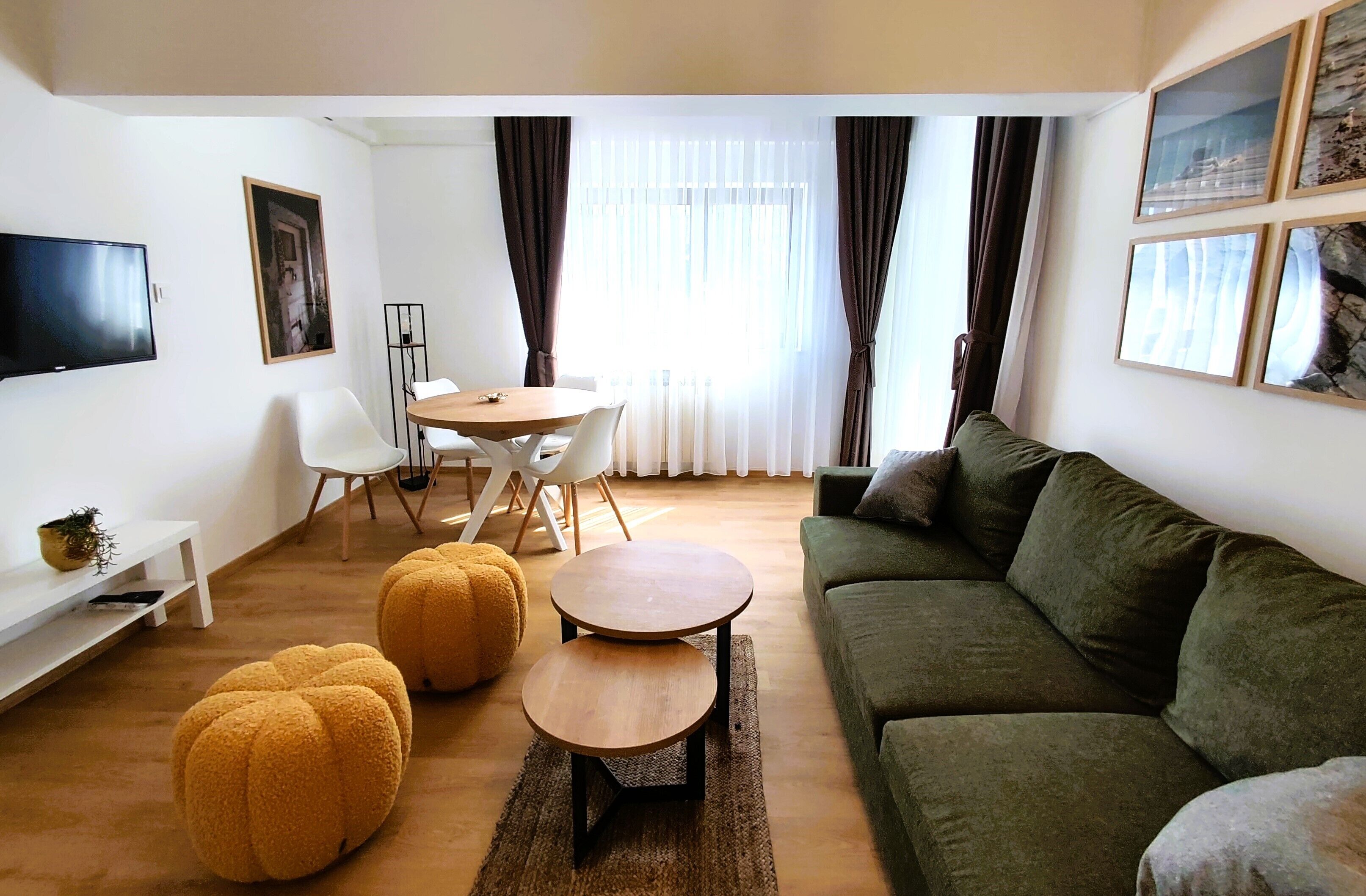 Una sala de estar moderna con un sofá verde, una mesa de centro de madera y una mesa redonda de comedor con sillas. Hay un televisor de pantalla plana montado en la pared, un estante blanco pequeño con una planta y un ventanal con cortinas.