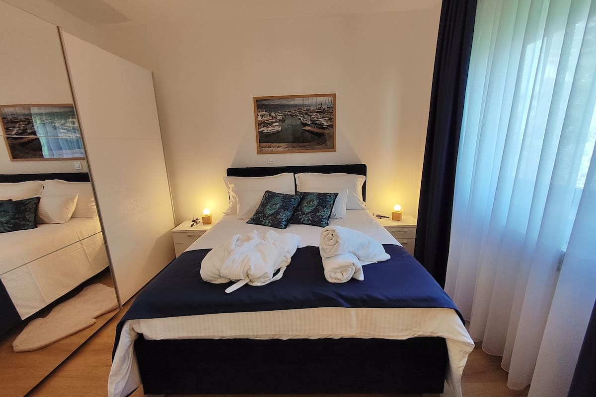Une chambre d’hôtel avec un grand lit, deux tables de chevet avec des lampes, un tableau au-dessus du lit et une vue sur la mer par la fenêtre.