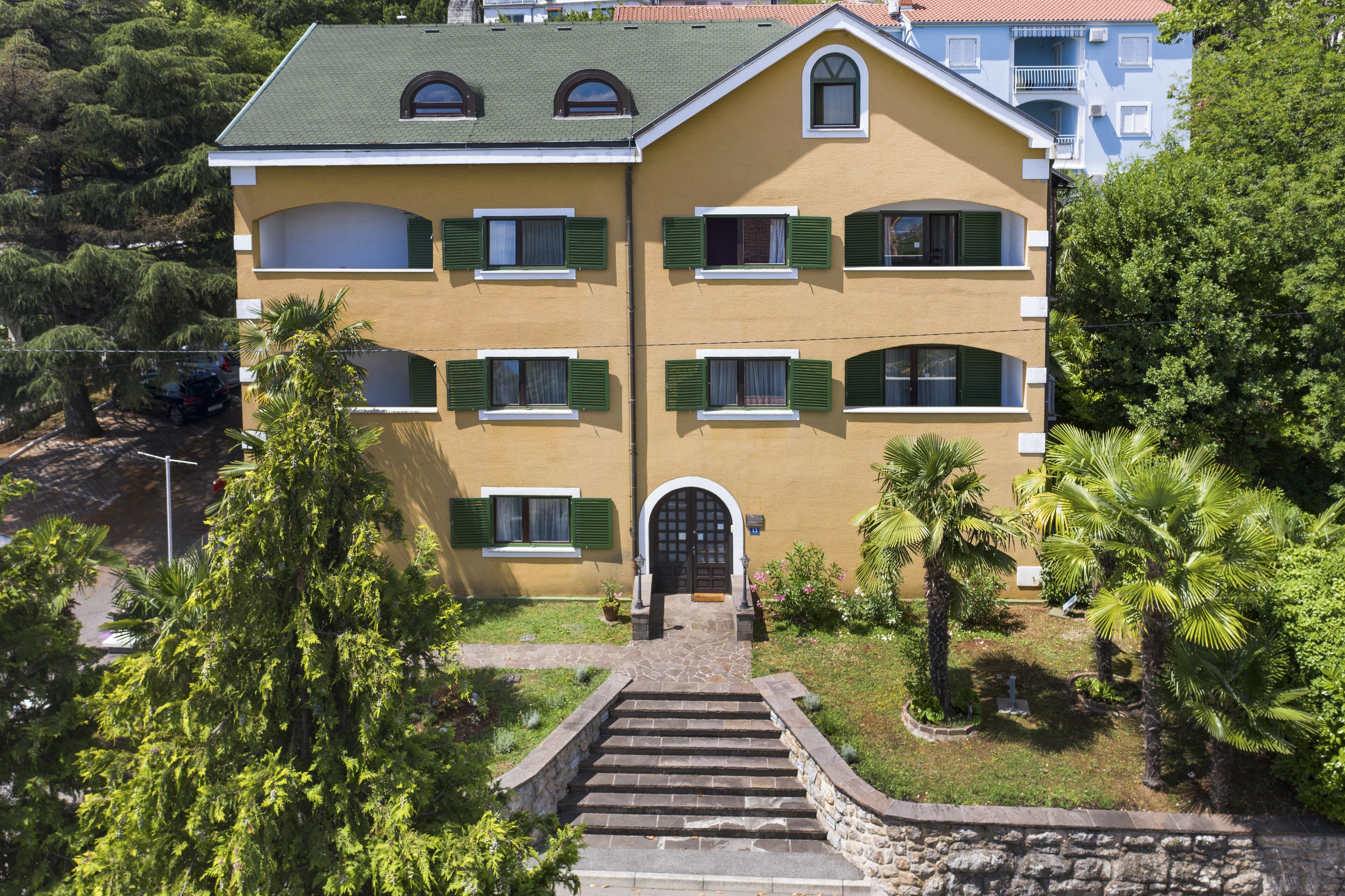 A multi-story building with a green roof, multiple windows with green shutters, and a central entrance with steps leading up to it.