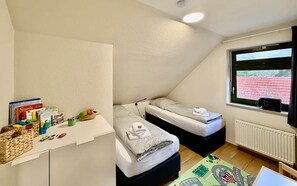 A small, triangular room with two beds, a white desk with books and toys, and a window with a view of a red fence. - Frisch Renoviertes, Sehr Modernes Ferienhaus für die Ganze Familie! (Varel)