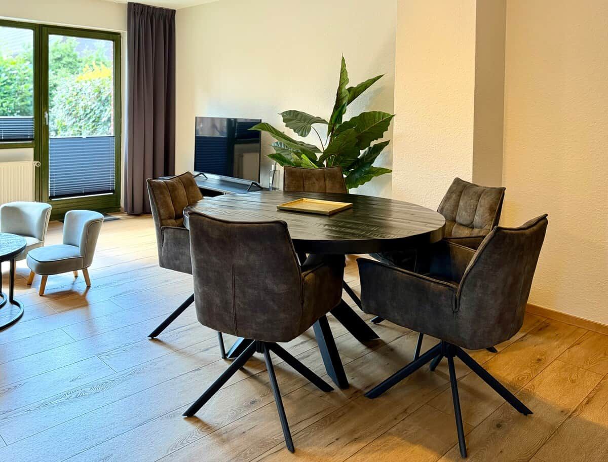A modern dining area with a round table and six chairs, a potted plant, and a view of the outdoors through a window.