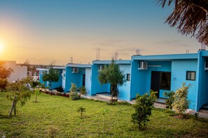 A row of blue buildings with air conditioning units, surrounded by small trees and grass. - Praia De Tofo (Tofo)