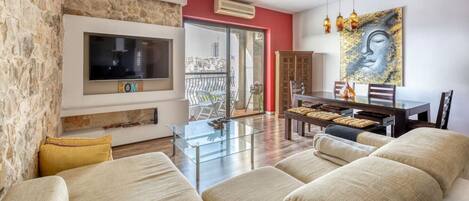 Una sala de estar con un sofá, una mesa de centro de vidrio, un televisor montado en una pared de piedra, y un comedor con una mesa y sillas.