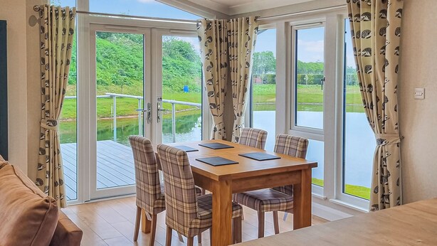 Un espace repas comprenant une table et des chaises, de grandes fenêtres avec des rideaux, et une vue sur la verdure et l’eau.