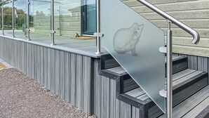 Un bâtiment moderne avec une façade vitrée, un balcon en verre, une rambarde en métal et une terrasse en bois.