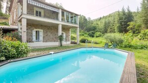 Una casa de piedra de dos plantas con piscina, rodeada de exuberante vegetación y colinas.