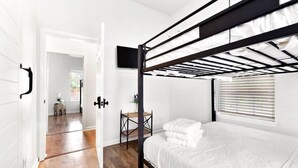 Habitación con literas, un televisor montado encima, una mesita con una planta y una ventana con persianas.