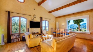Un salón amplio con ventanas grandes, un televisor de pantalla plana, una zona de comedor y una estantería.