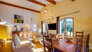 Un comedor con una mesa de madera, sillas y un televisor. Hay ventanas con cortinas, un sofá y un armario.