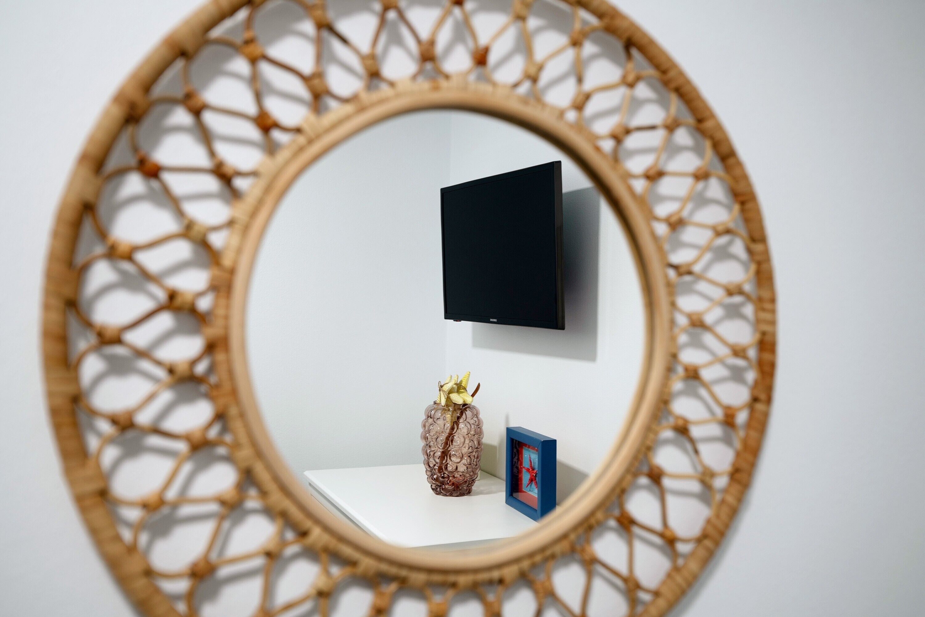 A round mirror with a geometric pattern, a flat-screen TV mounted on the wall, a small decorative vase, and a picture frame on a white shelf.