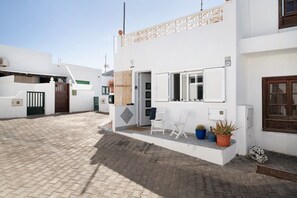 Un edificio blanco con un patio empedrado, una puerta de madera y un pequeño rincón con sillas y plantas en macetas.