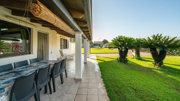 An outdoor patio with a dining table and chairs, a covered area with a ceiling, and a lawn with palm trees. - Vacation Home "Casa Silvia" with Private Terrace & Wi-Fi (Calasetta)