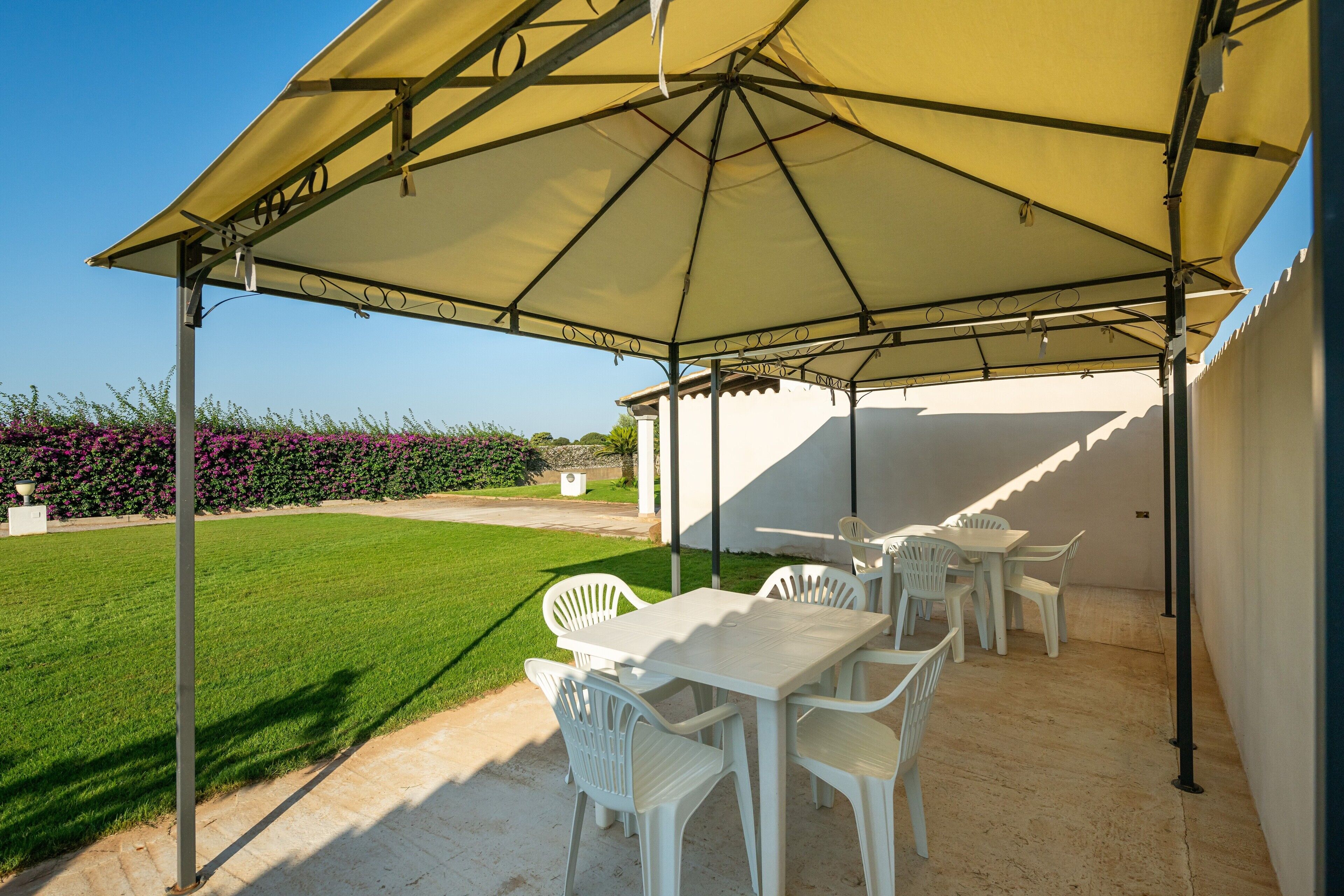 Un pati exterior amb taula i cadires sota un parasol gran, envoltat d'un jardí ben cuidat i una paret blanca.
