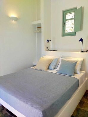 Ein Bett mit grauer Bettdecke, zwei Nachttische mit Lampen, ein Fenster mit grünem Rahmen und eine Wandleuchte.