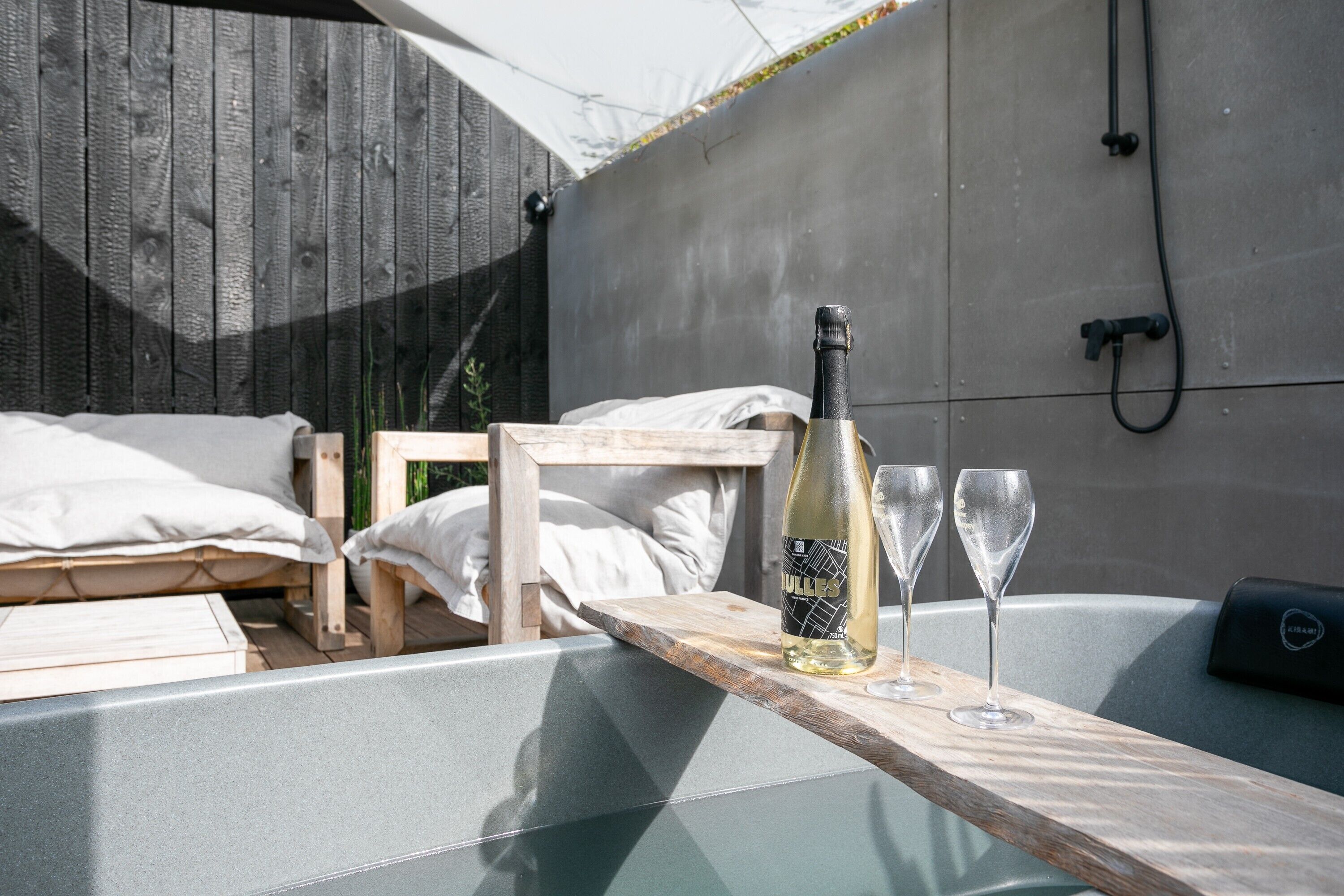 Un banc en bois avec une bouteille de vin et deux verres posés sur une table, un lit avec une literie blanche et, au fond, un mur en béton avec un tuyau.