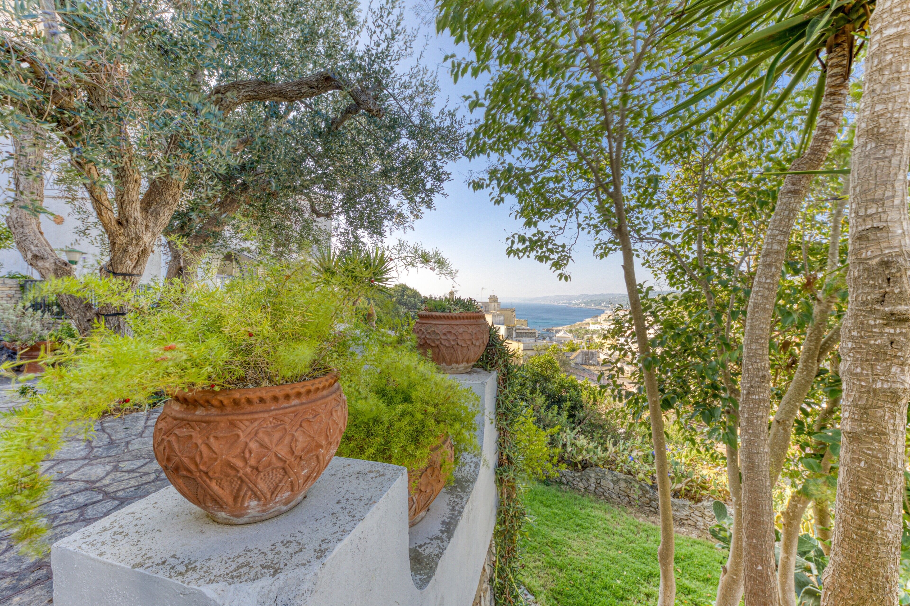 Un jardín con plantas en macetas y árboles, un sendero de piedra y vistas al mar.