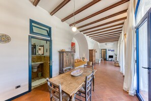 A spacious dining area with a wooden table and chairs, a tiled floor, and a high ceiling with exposed beams.