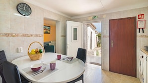 A dining area with a round table, chairs, a clock, a basket of fruit, and a door leading outside.