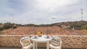 An outdoor patio with a table set for two, a basket of fruits, and a view of terracotta-tiled roofs.