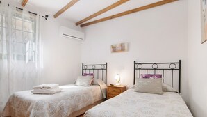 A bedroom with two beds, a nightstand with a lamp, a window with sheer curtains, and an air conditioning unit on the wall. - Holiday Home "El Llanete" with Mountain Views, Shared Pool & Wi-Fi (Vega de San Mateo)