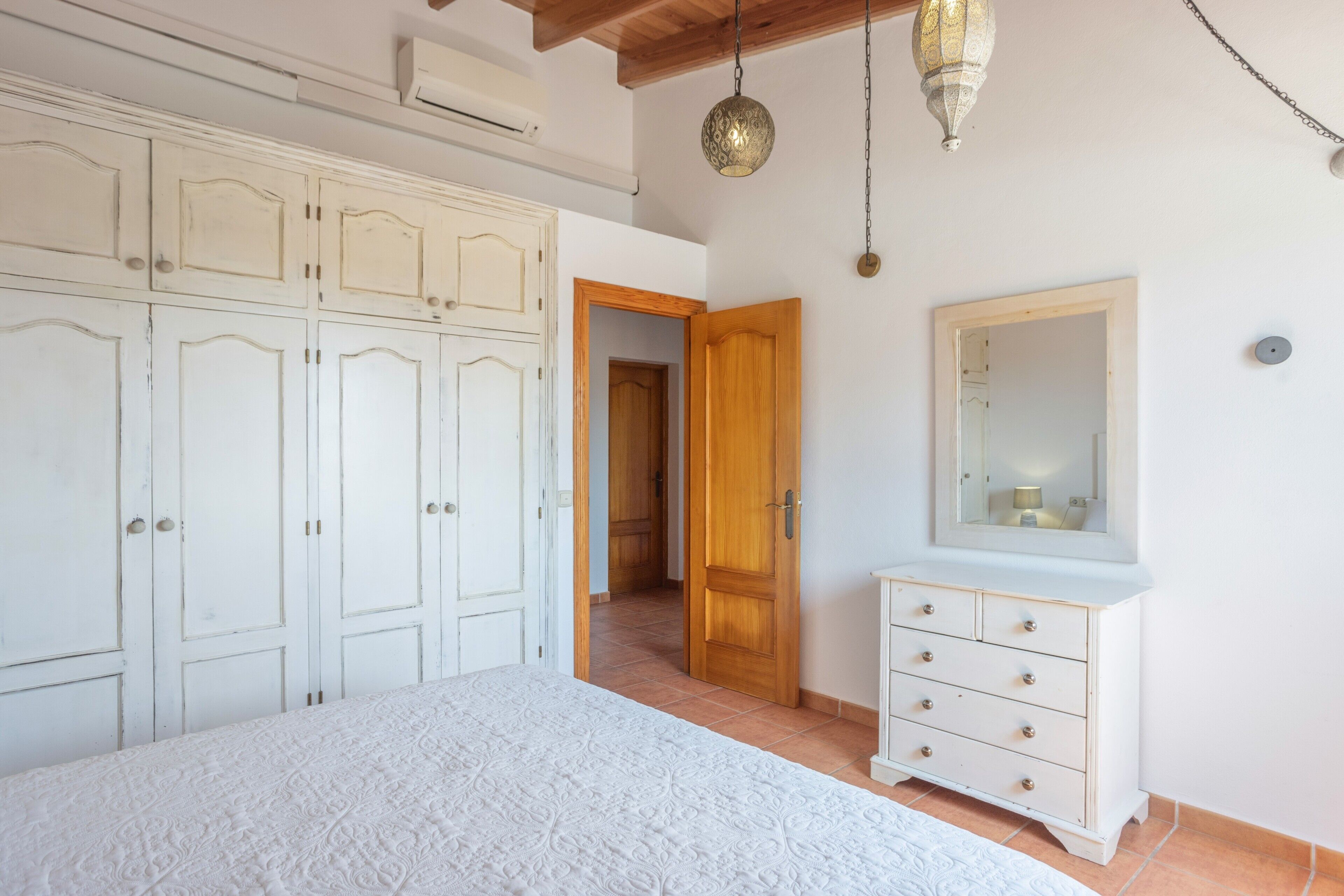 Un dormitorio con cama, armario, cómoda, espejo y lámpara colgante.