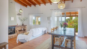 A spacious living area with a stone fireplace, a sofa, a dining table with chairs, and a large window with a view of greenery. - Holiday Home 'Can Juan Pedro' with Sea View, Wi-Fi and Air Conditioning (Formentera)