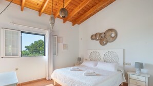 A bedroom with a wooden ceiling, a bed with white linens, a nightstand, a window with curtains, and a view of trees outside. - Holiday Home 'Can Juan Pedro' with Sea View, Wi-Fi and Air Conditioning (Formentera)