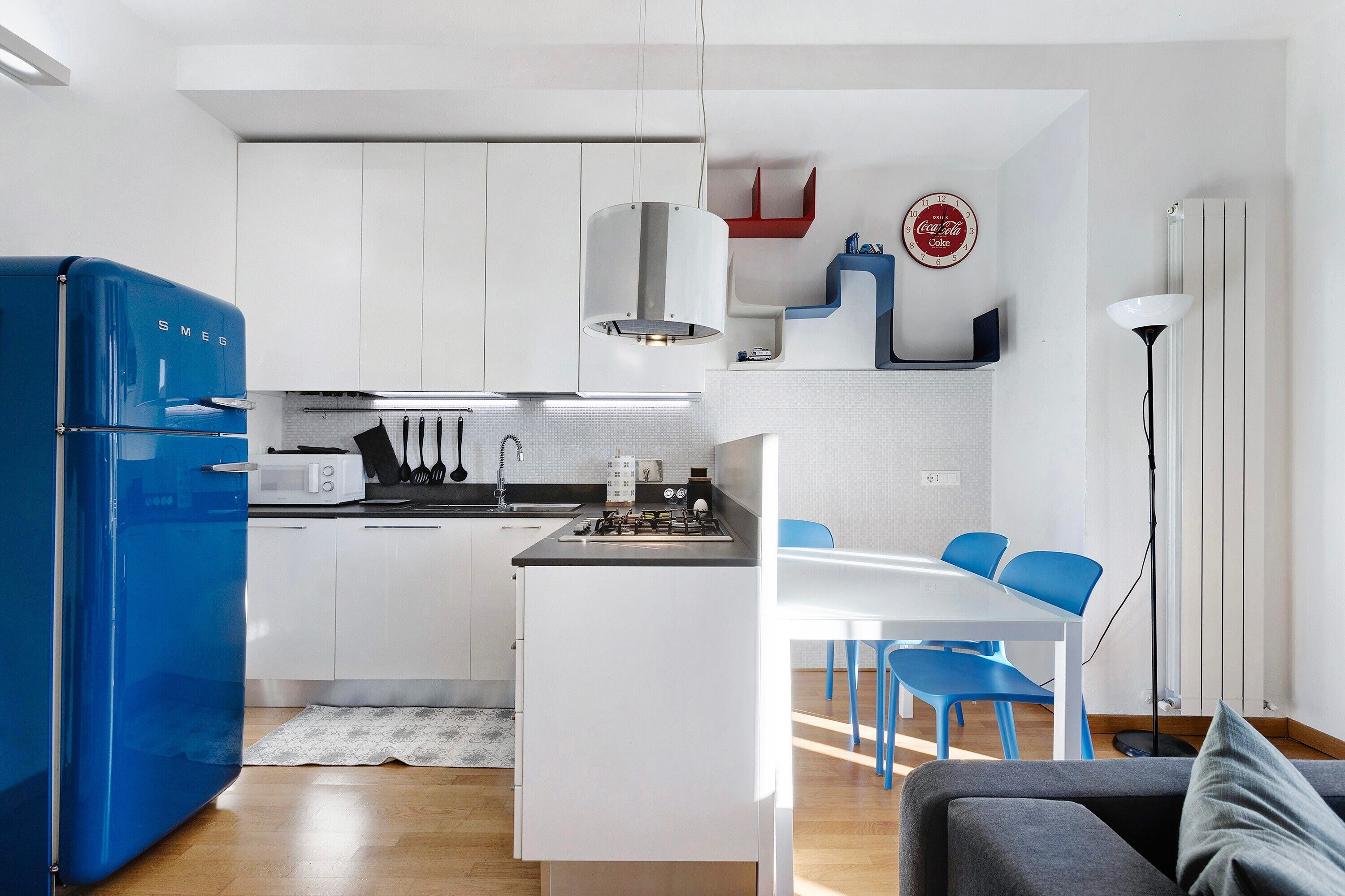 A compact kitchen with white cabinets, a blue refrigerator, a dining area with blue chairs, and a floor lamp.
